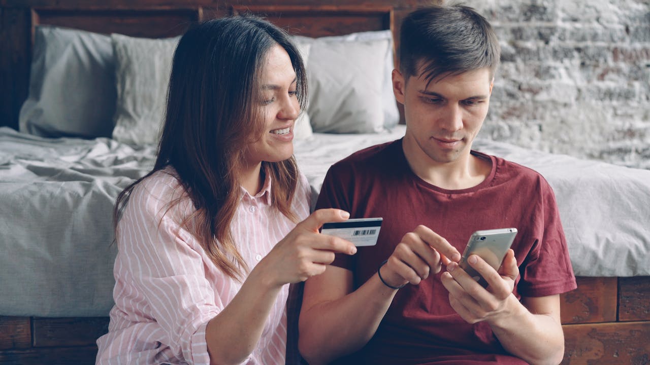 gallery-1 A couple sits on a bed shopping online using a smartphone and credit card in a cozy indoor setting.