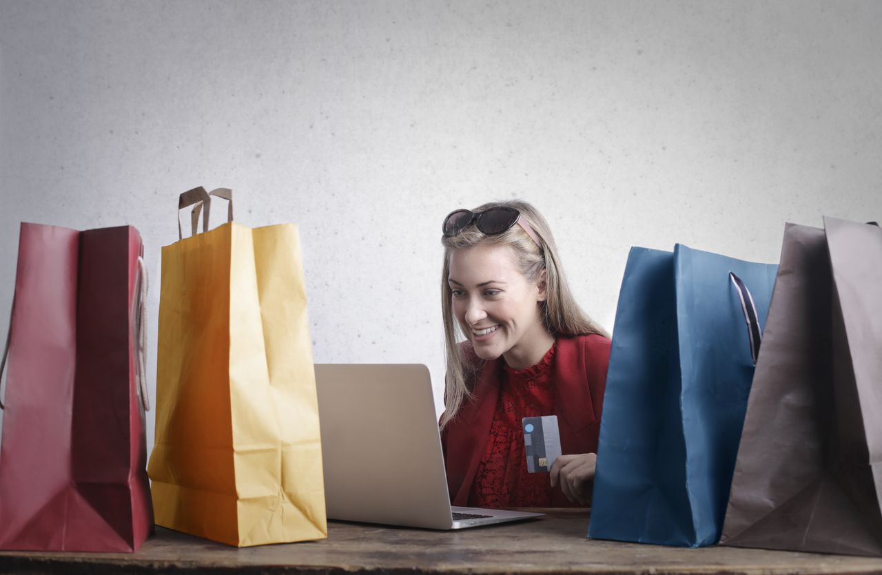 services-03 Cheerful young female with credit card in hand using laptop and enjoying shopping online while sitting at table with various colorful shopping bags
