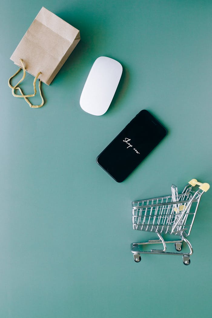 gallery-6 Top view of a smartphone, mouse, paper bag, and mini shopping cart on a green background, symbolizing e-commerce.