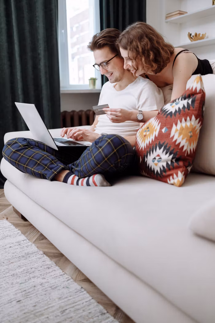 services-02 Happy couple sitting on a couch, using a laptop for online shopping with a credit card in hand.