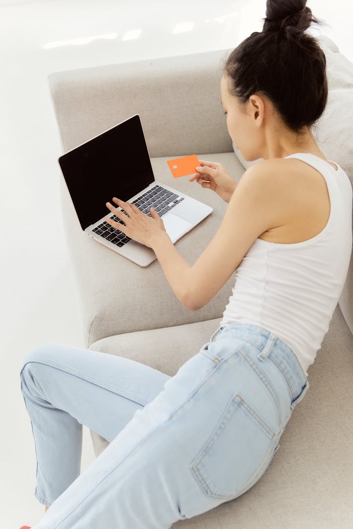 gallery-4 Young woman using a laptop and credit card for online shopping while lounging on a sofa.